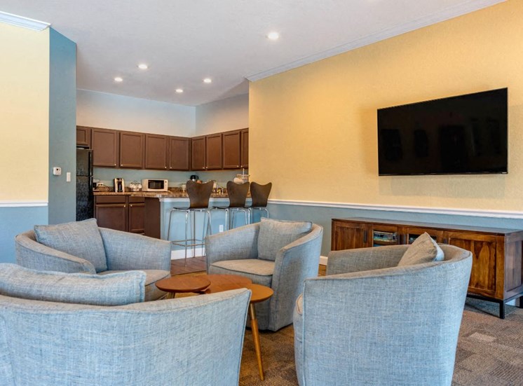 Resident Lounge Area with Mounted TV Grey Chairs Around Coffee Table and Clubhouse Kitchen with Brown Cabinets